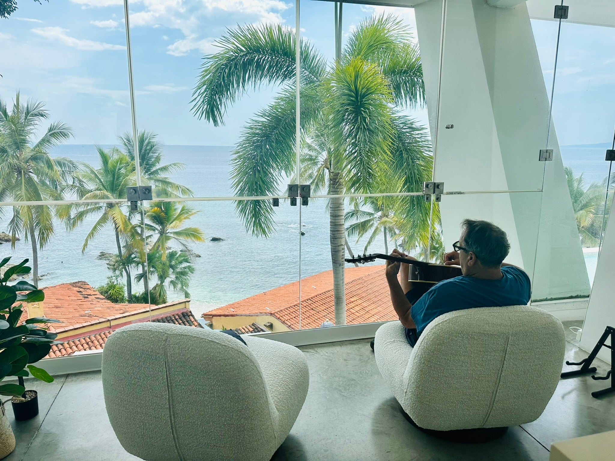 A man sits in a modern white lounge chair, playing a black guitar near a large floor-to-ceiling window with a scenic view of palm trees, the ocean, and blue sky.