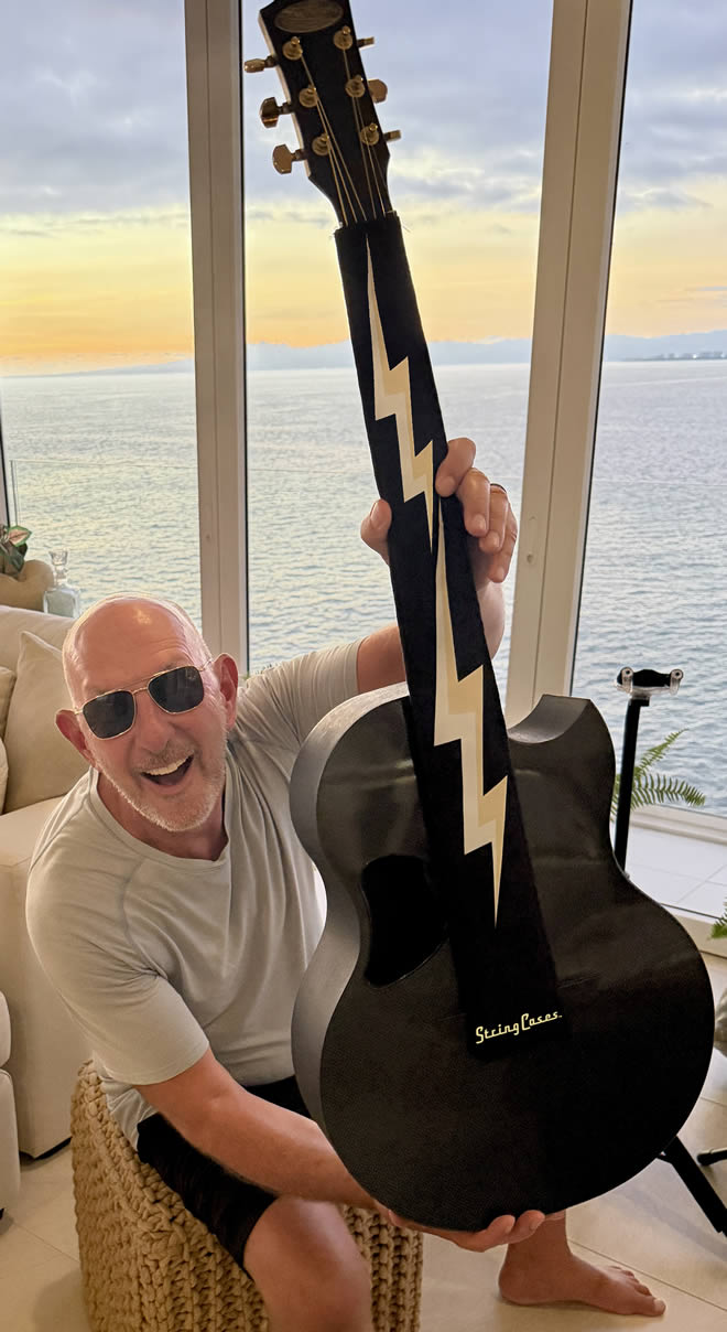 Man holding a black electric guitar with lightning bolt string case design indoors, ocean view in the background

Details